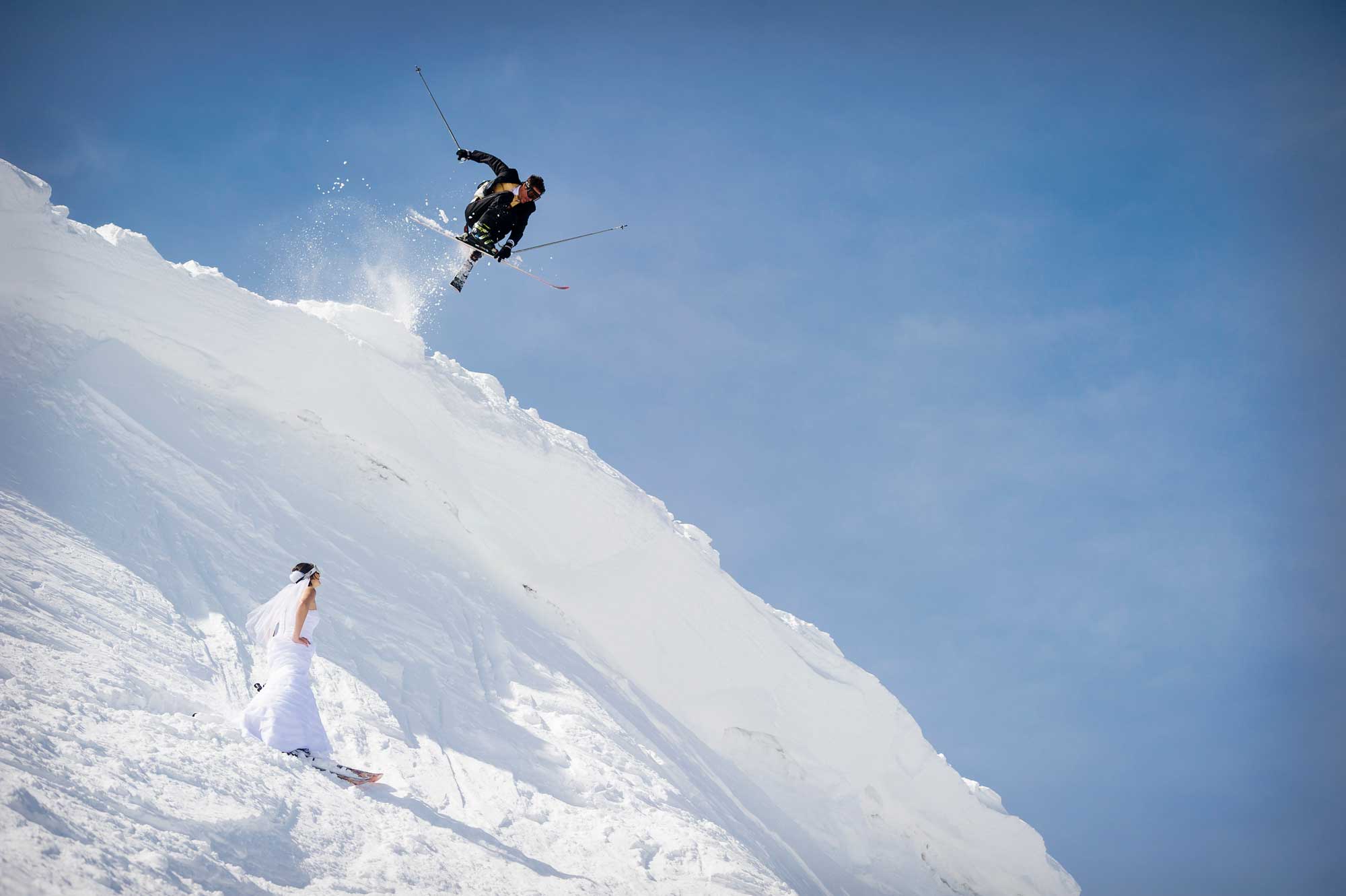 Adventure ski wedding photography of groom jumping over bride on skis